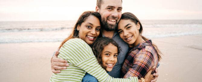 A laughing family of four sharing a group hug on a beach, featuring a mother, father, a preteen daughter, and a younger child