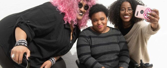 A tight crop of three Black and disabled friends (a non-binary person with a cane and tangle stim toy, a non-binary person sitting in a power wheelchair, and an invisibly disabled femme) smiling and taking a cell phone selfie together.