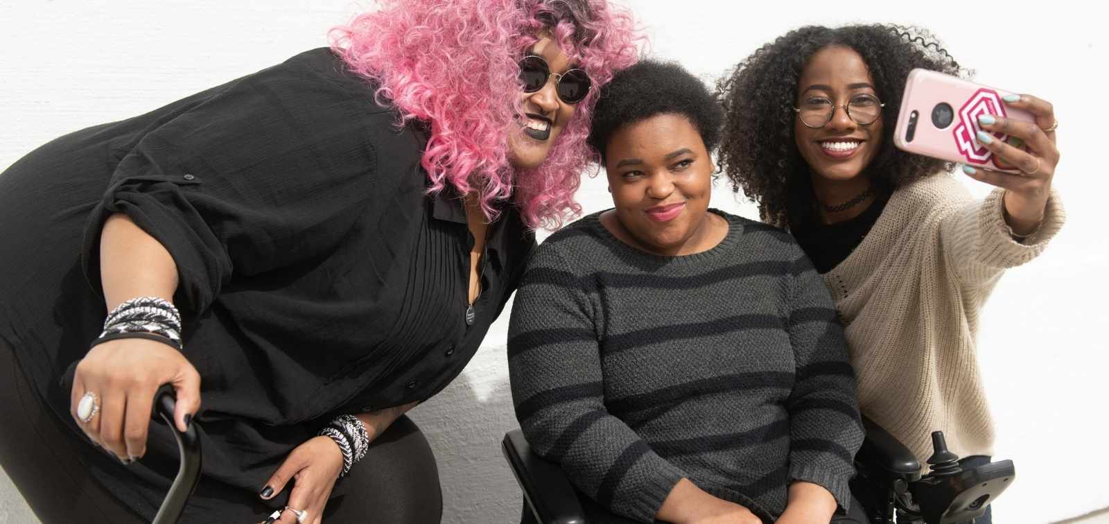 A tight crop of three Black and disabled friends (a non-binary person with a cane and tangle stim toy, a non-binary person sitting in a power wheelchair, and an invisibly disabled femme) smiling and taking a cell phone selfie together.